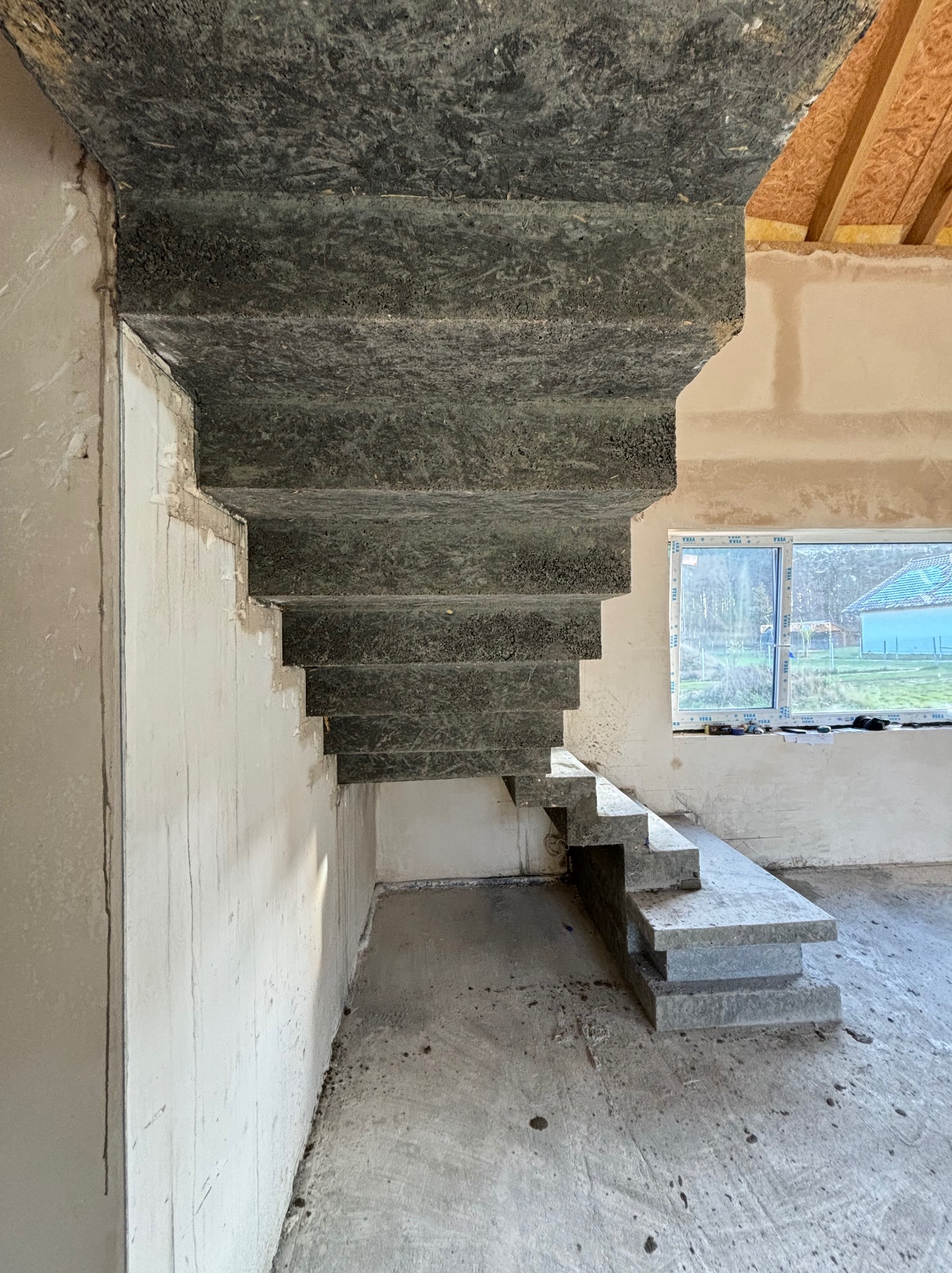Surowe, betonowe schody w trakcie budowy w jasnym, niedokończonym wnętrzu z widocznym oknem i elementami konstrukcji dachu.