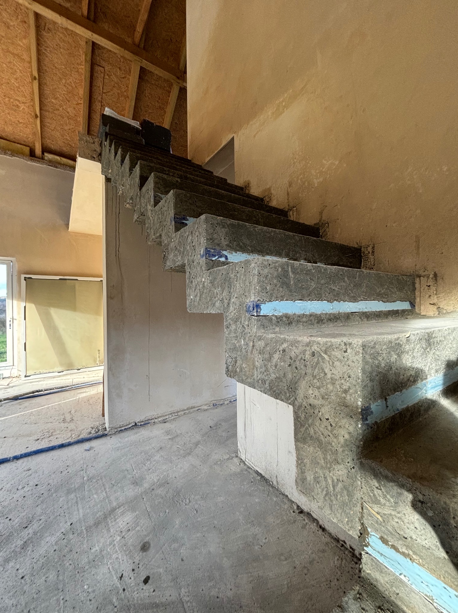 Surowe, niewykończone schody z betonu komórkowego z niebieską taśmą zabezpieczającą krawędzie, widoczne w surowym wnętrzu budynku w trakcie budowy.