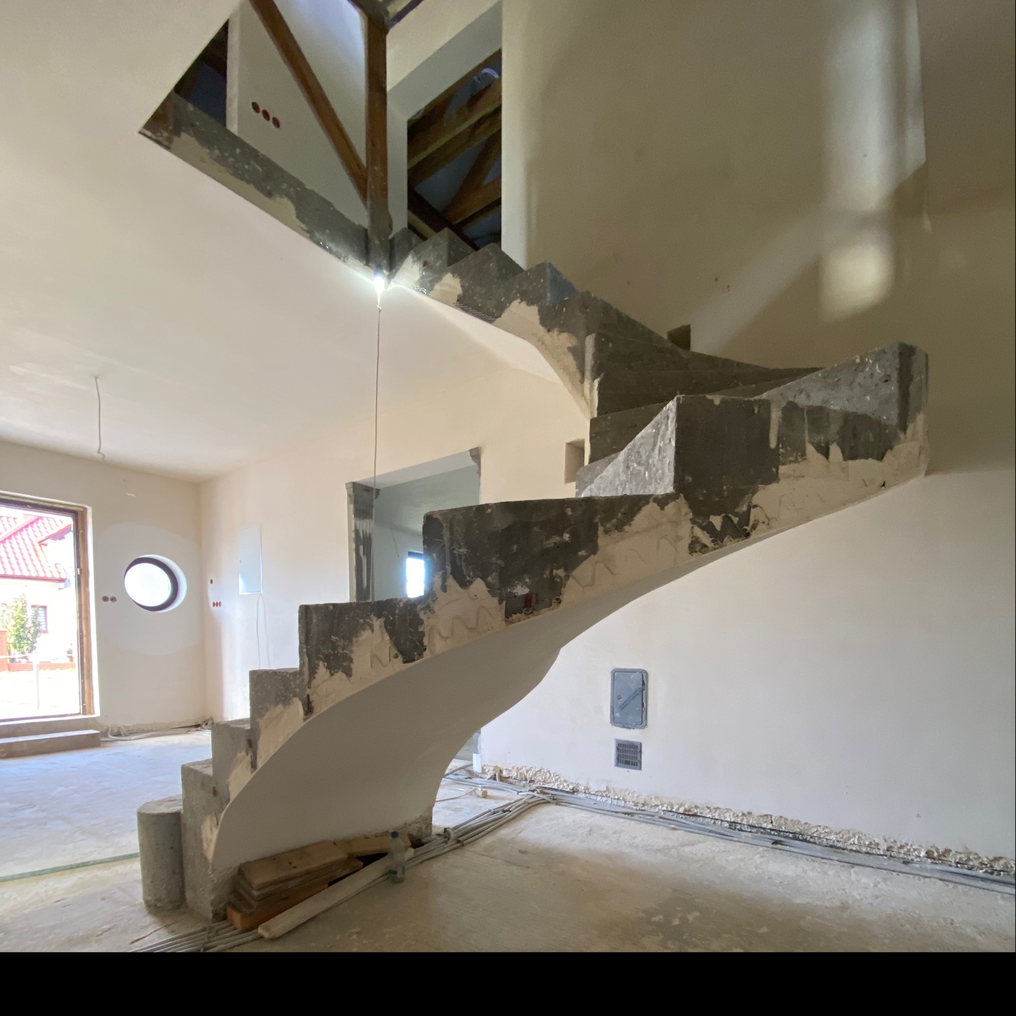 Wnętrze w trakcie wykańczania z spiralnymi schodami w surowym betonie, widoczne instalacje podłogowe i otwór w stropie z elementami drewnianymi.