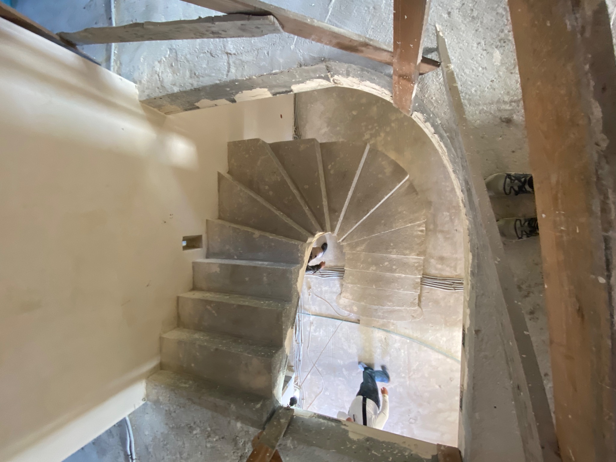 Spiralne schody w surowym stanie, widok z góry podczas prac wykończeniowych, widoczne kable i fragmenty rusztowania, osoba w roboczym ubraniu na dole.