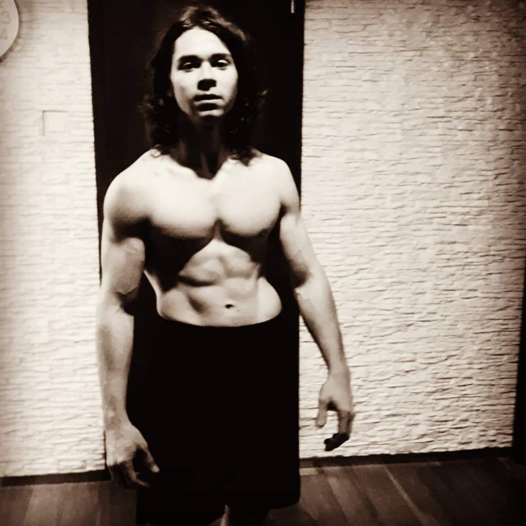 Mężczyzna z długimi włosami, bez koszulki, prezentuje wyrzeźbioną sylwetkę w sepii, na tle ściany z dekoracyjnej cegły i ciemnych drzwi.