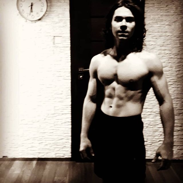 Mężczyzna z długimi włosami prezentuje wyrzeźbioną sylwetkę w czarno-białym ujęciu, widoczny zegar na ścianie i zamknięte drzwi w tle.