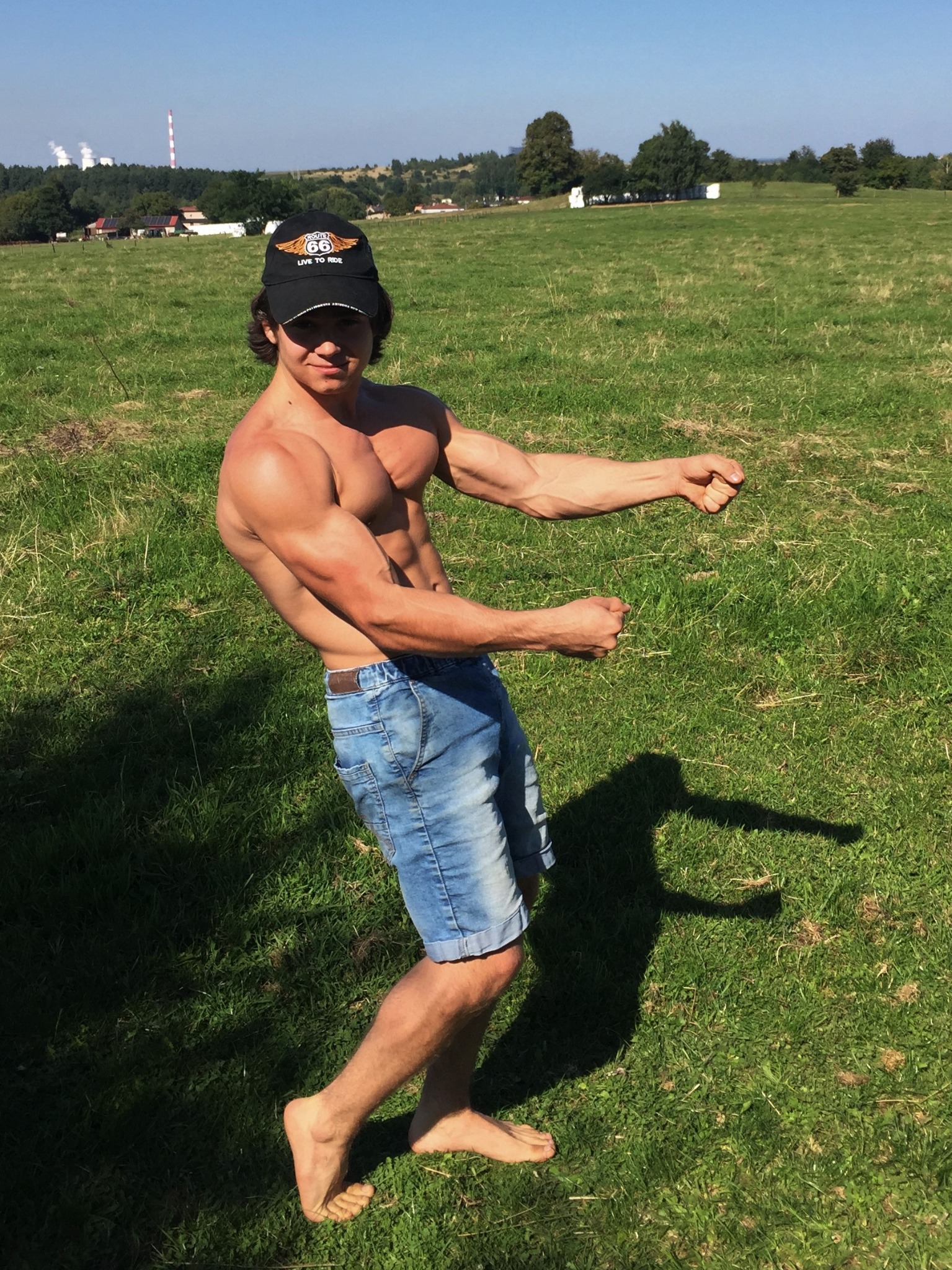 Młody, opalony mężczyzna bez koszulki, w dżinsowych szortach i czapce z daszkiem, prezentuje swoją muskulaturę na zielonej łące w słoneczny dzień.