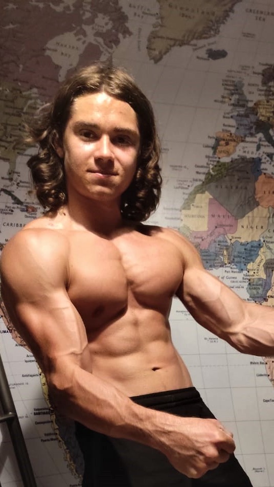 Młody mężczyzna z umięśnioną sylwetką, pozuje na tle mapy świata, prezentując napięte bicepsy i zarysowane mięśnie brzucha.