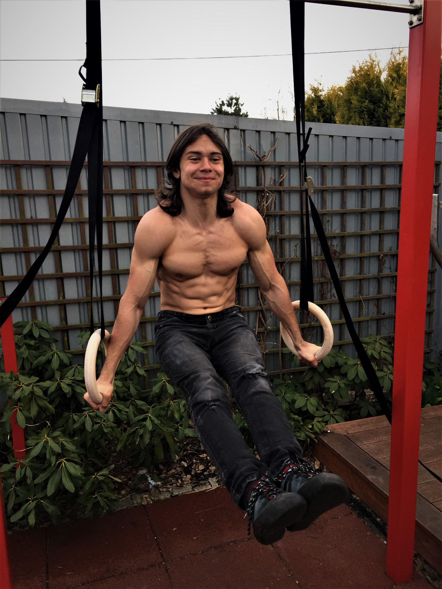 Uśmiechnięty, umięśniony mężczyzna z długimi włosami ćwiczy na drewnianych kółkach gimnastycznych na zewnątrz, w tle szary metalowy płot i zielone krzewy.