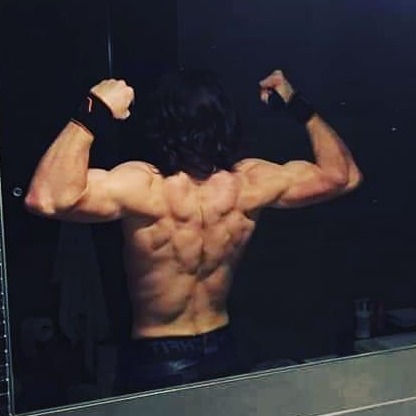 Mężczyzna z długimi włosami, odwrócony tyłem, prezentuje rozbudowane mięśnie pleców i ramion, nosząc czarne opaski na nadgarstkach, widoczny w lustrze łazienki o ciemnym otoczeniu.