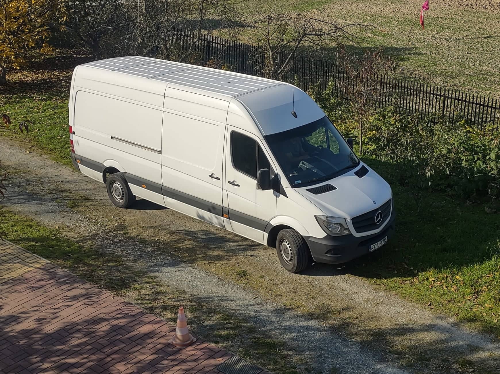 Biały samochód dostawczy marki Mercedes-Benz zaparkowany na żwirowej drodze, widziany z góry, z pomarańczowym pachołkiem na pierwszym planie.