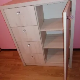 Jasna komoda z trzema szufladami i półkami, otwarte drzwiczki z ozdobnym kryształowym uchwytem, widoczne na tle różowej ściany i drewnianej podłogi.