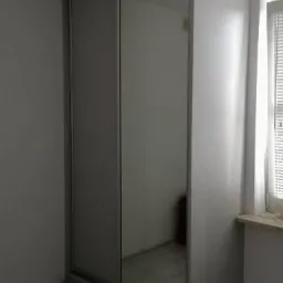 Szara szafa przesuwna z lustrem w białym pokoju, widok z boku, odbicie okna w lustrze.