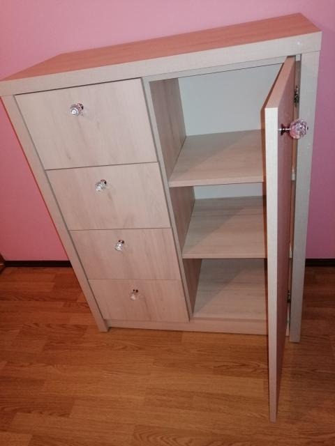 Jasna komoda z trzema szufladami i półkami, otwarte drzwiczki z ozdobnym kryształowym uchwytem, widoczne na tle różowej ściany i drewnianej podłogi.