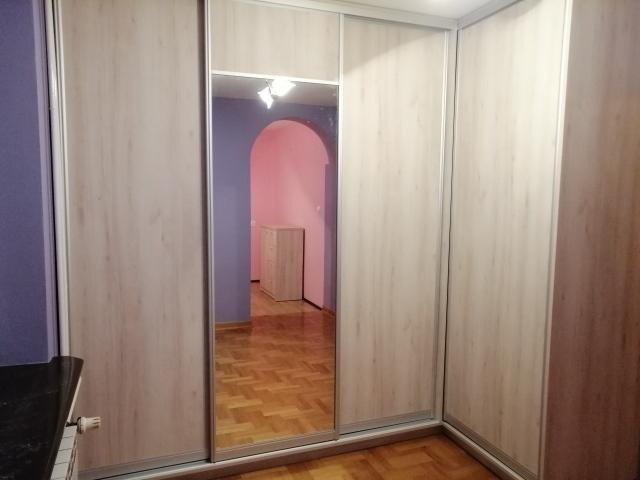 Jasna, narożna szafa z przesuwnymi drzwiami, z lustrem w jednych drzwiach, odbijającym widok na pokój z parkietem i łukowym przejściem.