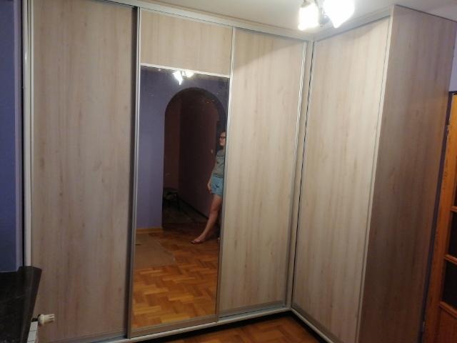 Jasna szafa wnękowa z przesuwnymi drzwiami, jedno skrzydło z lustrem odbijającym wnętrze pokoju z widoczną osobą.