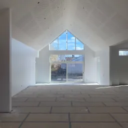 Jasne, przestronne wnętrze z wysokim sufitem wykończonym płytami z perforacją. Ściany białe, podłoga przygotowana do dalszych prac. Duże okno z widokiem na zewnątrz.