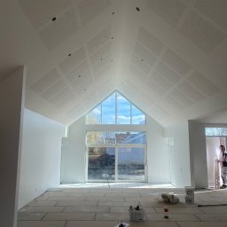 WS Wojciech Stempowski - Białe, wysokie wnętrze po malowaniu w Chojnie, z oknem szczytowym i perforowanym sufitem. Widoczne narzędzia malarskie i wiaderka z farbą. Minimalizm i jasność.
