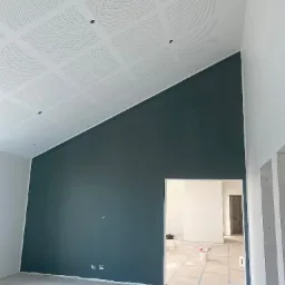 Pomieszczenie po malowaniu: ciemnozielona ściana pod skośnym, perforowanym sufitem, widok przez drzwi na dalszą część wnętrza w trakcie prac wykończeniowych.