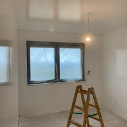 WS Wojciech Stempowski - Białe, świeżo pomalowane wnętrze pokoju z oknem zabezpieczonym folią malarską, drabina drewniana w rogu. Widoczne ślady farby na podłodze.