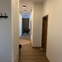 Długi korytarz z drewnianą podłogą i białymi ścianami, oświetlony rzędem okrągłych lamp sufitowych. W tle widoczna drewniana ściana i ławka.