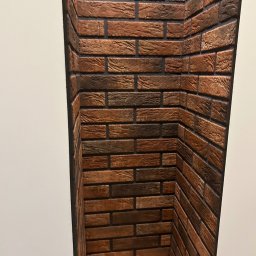 WS Wojciech Stempowski - Wnęka wykończona dekoracyjną cegłą w odcieniach brązu i grafitu, z drewnianą półką na dole. Widoczne na tle białej ściany.