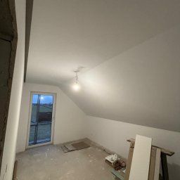 WS Wojciech Stempowski - Poddasze po malowaniu na biało, widoczne skosy, okno w szczycie, sufit oświetlony żarówką. Na podłodze resztki materiałów budowlanych i narzędzia.