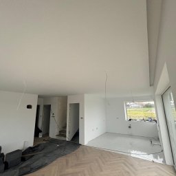 WS Wojciech Stempowski - Jasne wnętrze w trakcie wykończenia: gładkie, białe ściany, parkiet w jodełkę, widoczne instalacje i okno z widokiem na zieleń. Częściowo zakryte podłoże.