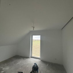 WS Wojciech Stempowski - Poddasze po szpachlowaniu i malowaniu na biało, widoczny odkurzacz przemysłowy i niedokończona instalacja elektryczna. W tle widok z okna na pole.