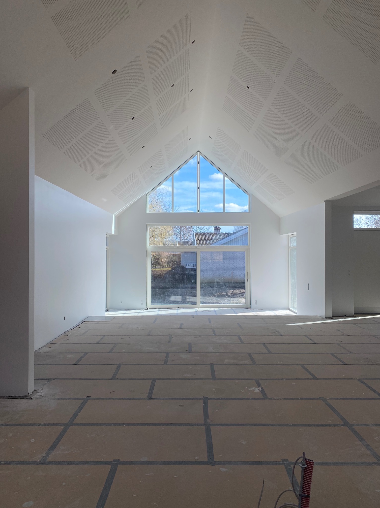 Jasne, przestronne wnętrze z wysokim sufitem wykończonym płytami z perforacją. Ściany białe, podłoga przygotowana do dalszych prac. Duże okno z widokiem na zewnątrz.