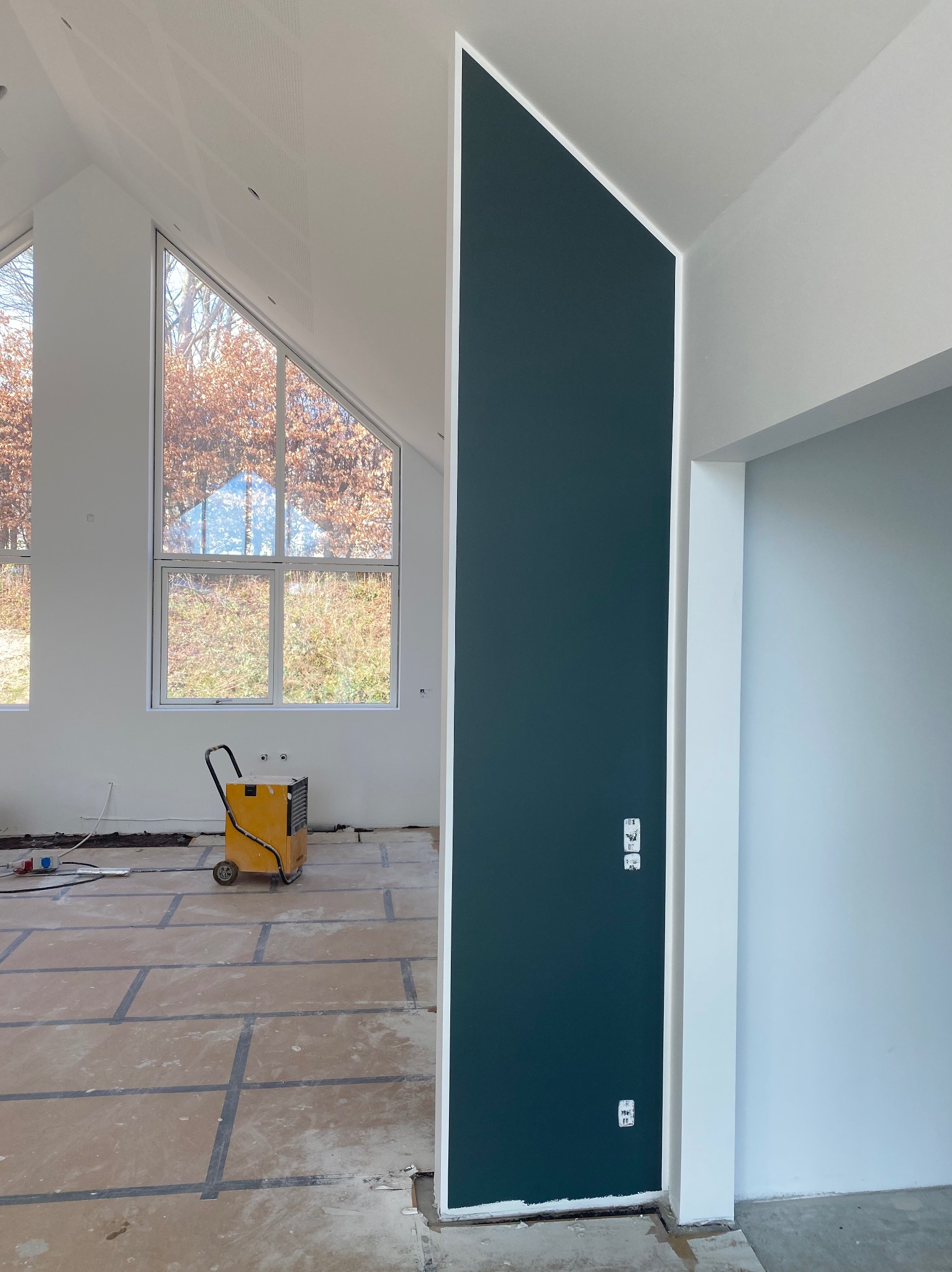 Wnętrze w trakcie wykańczania: pomalowana ściana w odcieniu butelkowej zieleni, widoczny osuszacz powietrza, duże okna i przygotowane podłoże pod posadzkę.