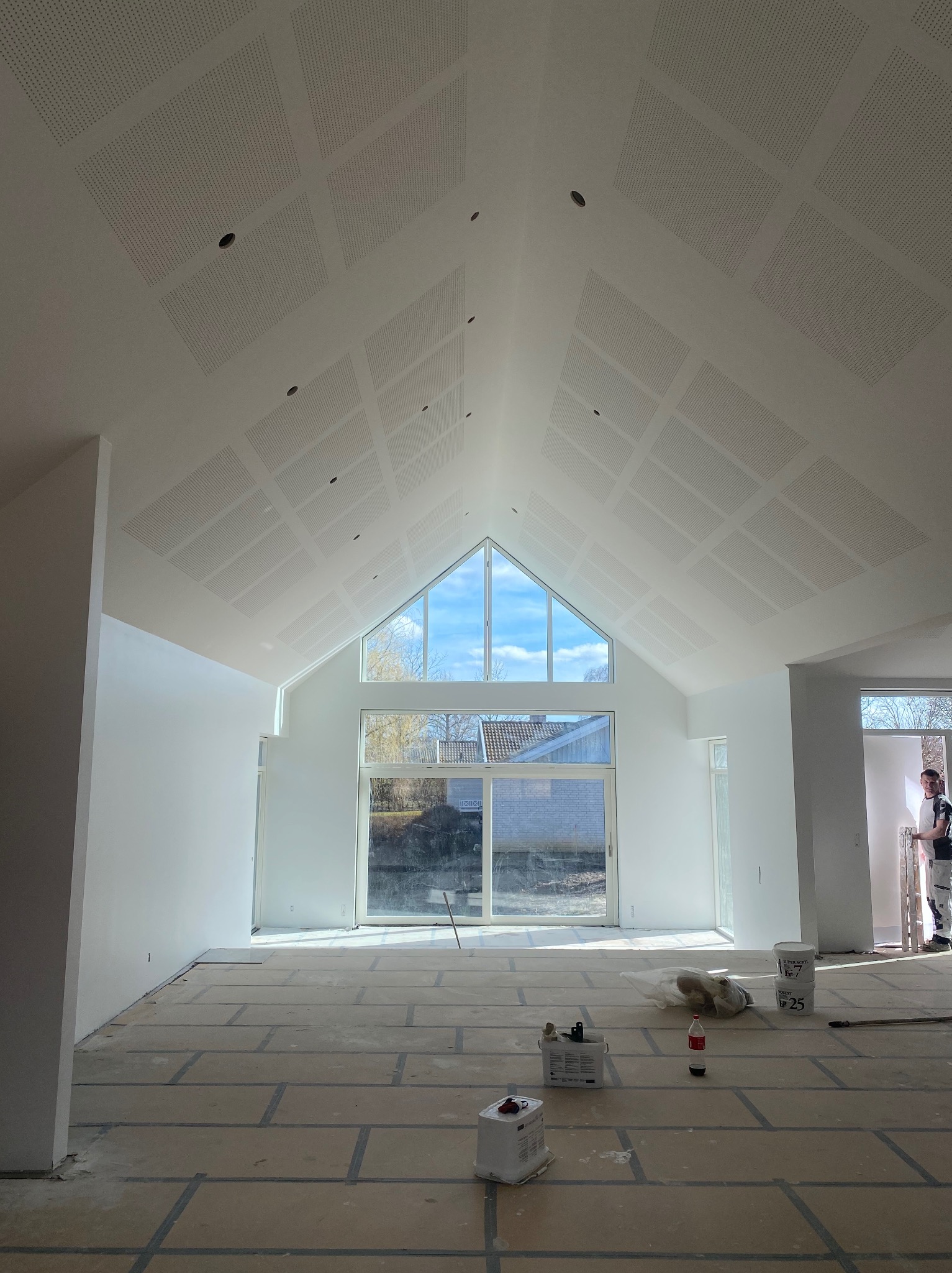 Białe, wysokie wnętrze po malowaniu w Chojnie, z oknem szczytowym i perforowanym sufitem. Widoczne narzędzia malarskie i wiaderka z farbą. Minimalizm i jasność.