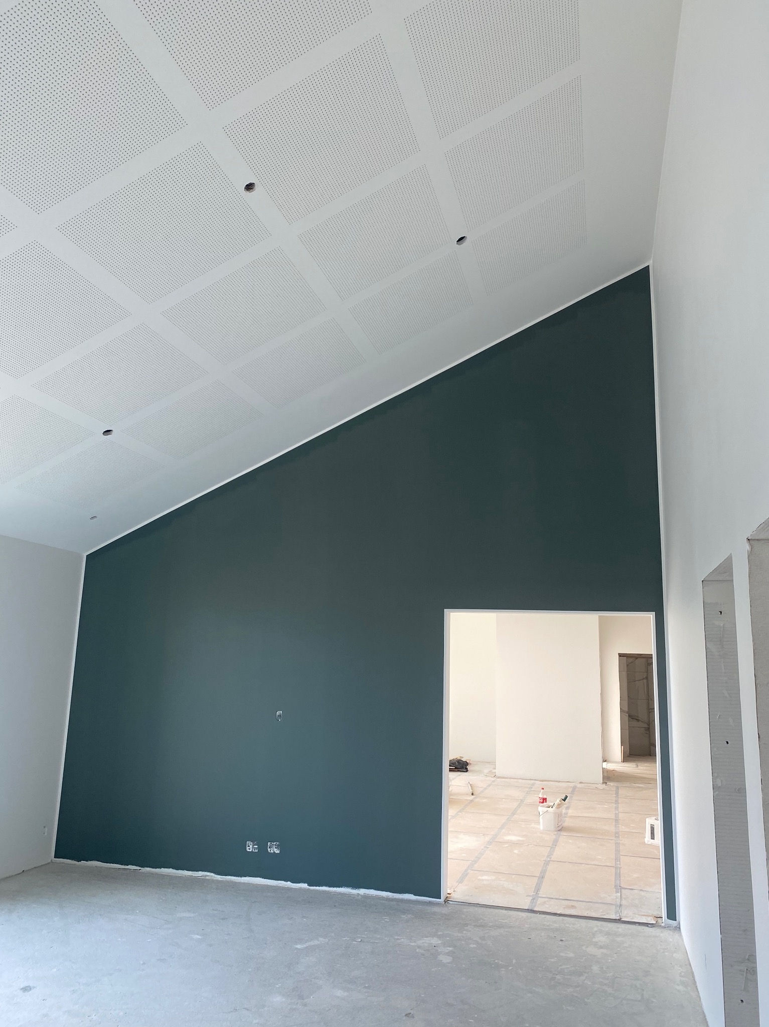 Pomieszczenie po malowaniu: ciemnozielona ściana pod skośnym, perforowanym sufitem, widok przez drzwi na dalszą część wnętrza w trakcie prac wykończeniowych.
