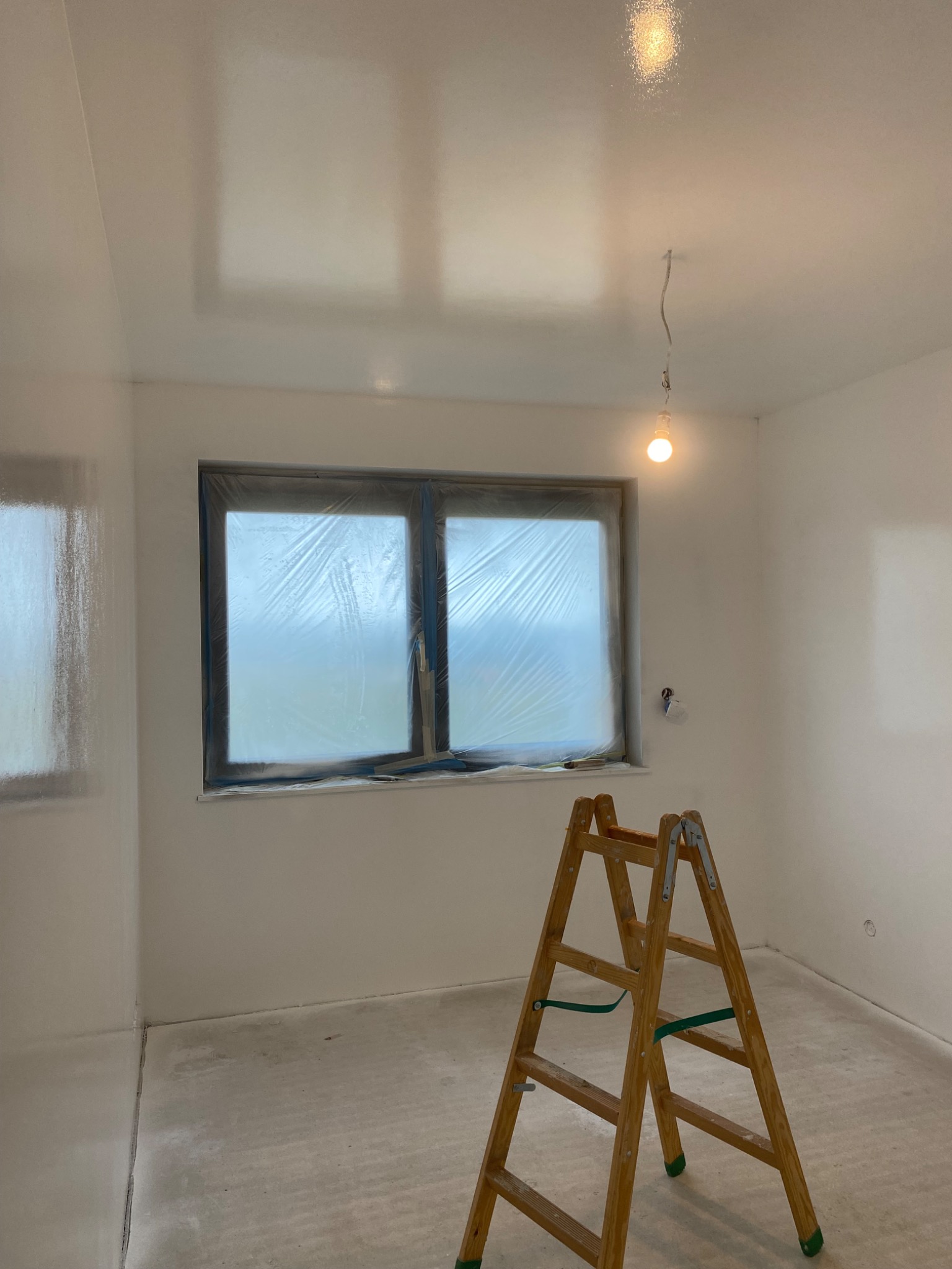 Białe, świeżo pomalowane wnętrze pokoju z oknem zabezpieczonym folią malarską, drabina drewniana w rogu. Widoczne ślady farby na podłodze.