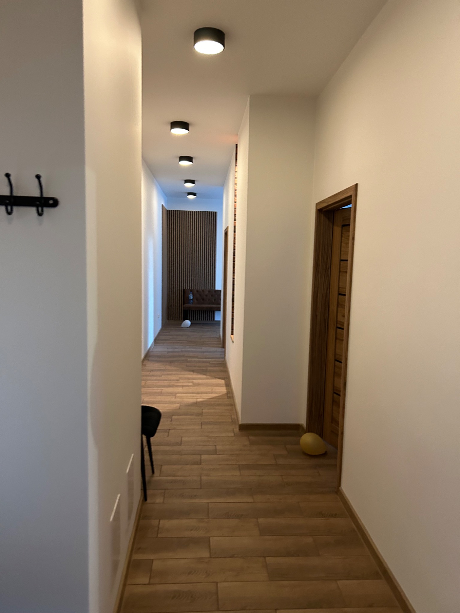 Długi korytarz z drewnianą podłogą i białymi ścianami, oświetlony rzędem okrągłych lamp sufitowych. W tle widoczna drewniana ściana i ławka.