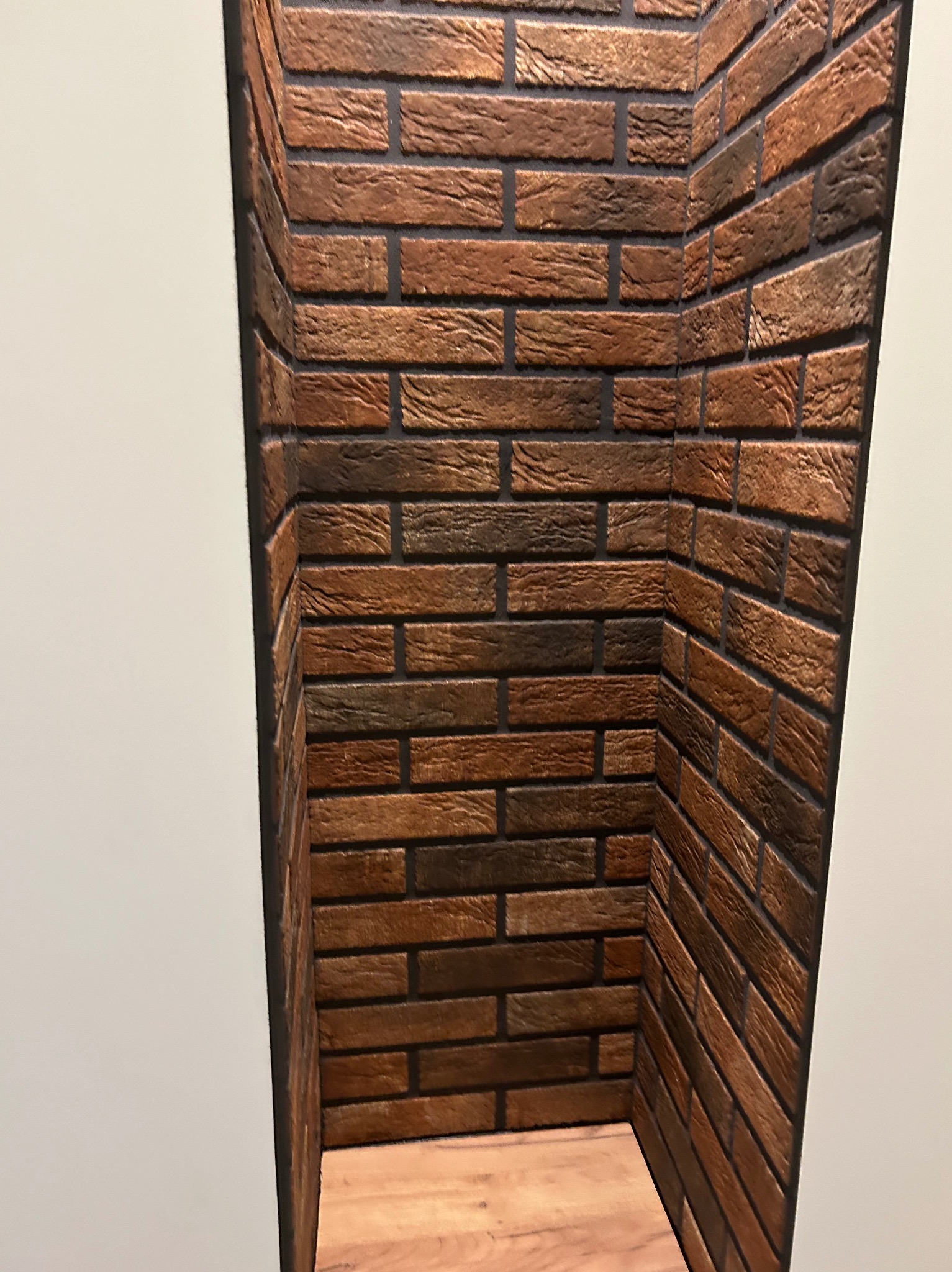 Wnęka wykończona dekoracyjną cegłą w odcieniach brązu i grafitu, z drewnianą półką na dole. Widoczne na tle białej ściany.