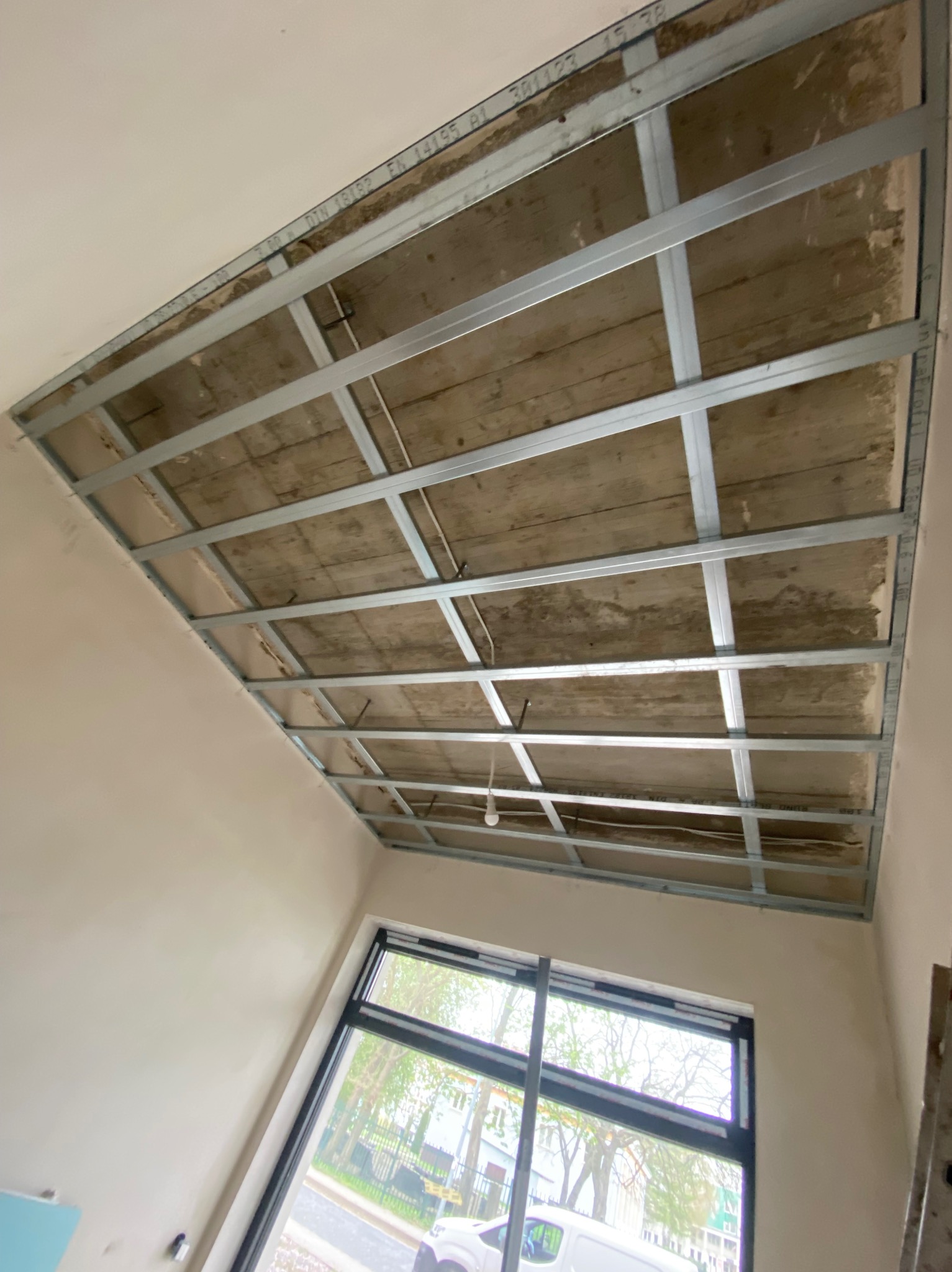 Montaż metalowego stelażu sufitu podwieszanego z płyty karton-gips w jasnym wnętrzu z oknem. Widoczne profile i instalacja elektryczna.