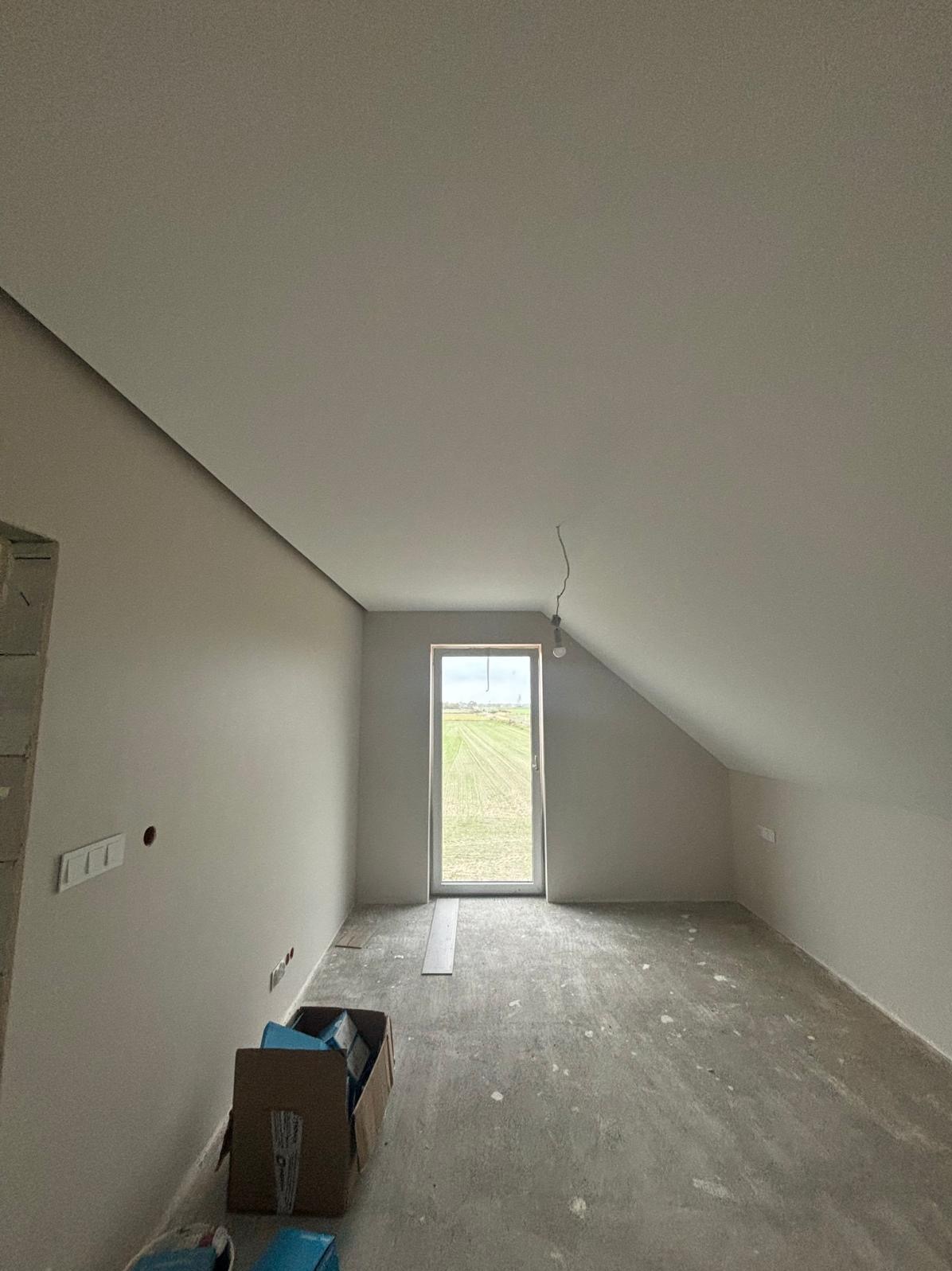 Poddasze po szpachlowaniu i malowaniu na neutralny kolor. Widoczne gniazdka elektryczne, okno z widokiem na pole i karton z materiałami budowlanymi.
