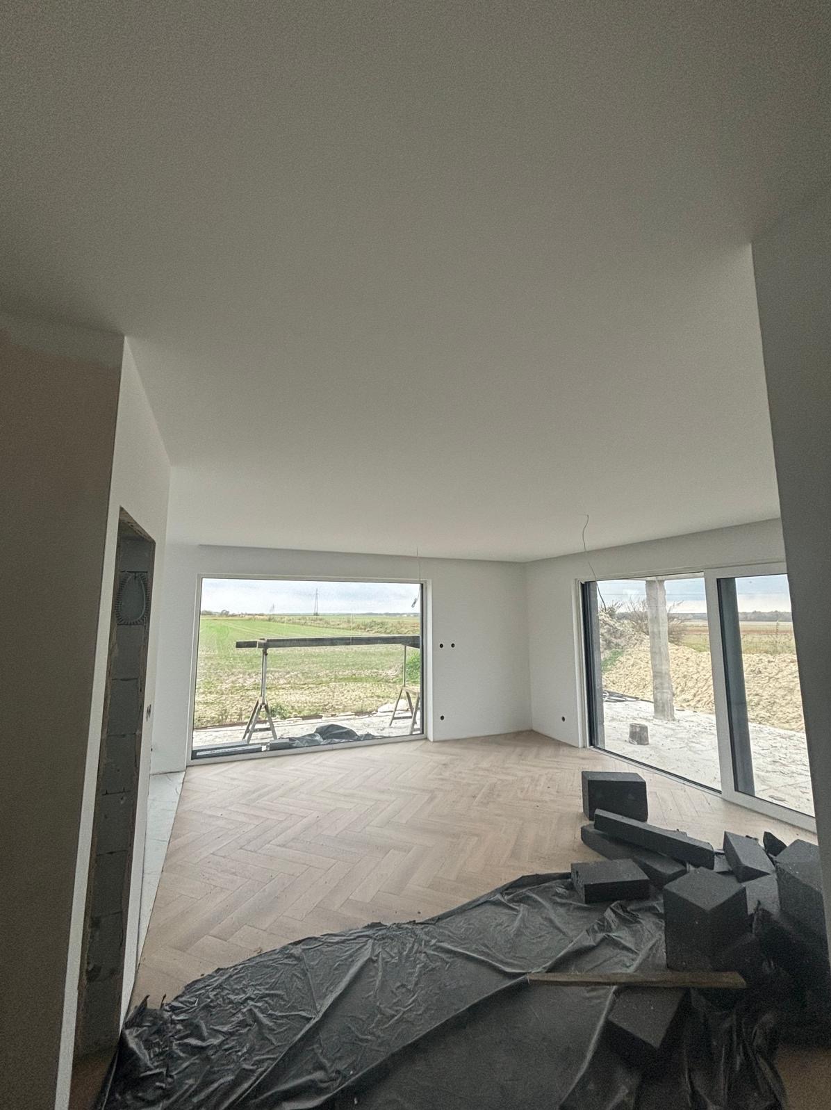 Wnętrze po szpachlowaniu i malowaniu, z parkietem w jodełkę i dużymi oknami. Widoczne materiały budowlane, co sugeruje trwający remont.