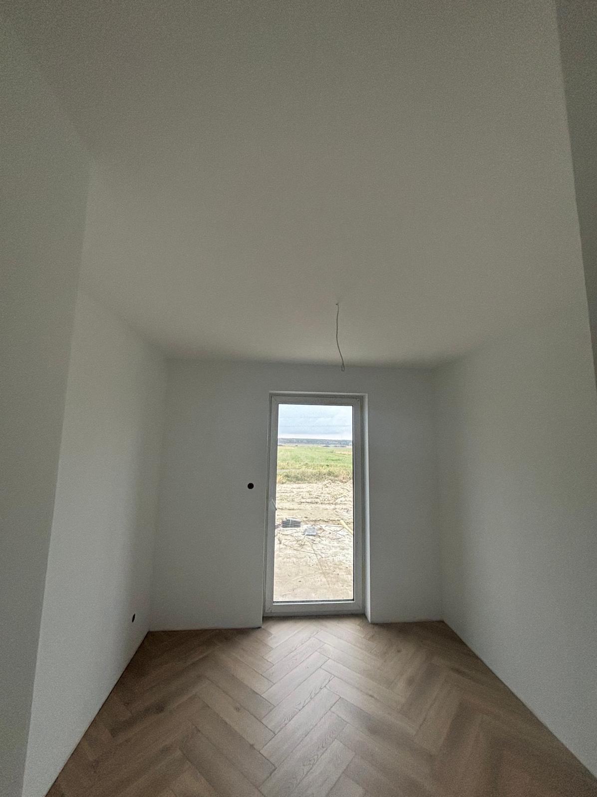 Pusty pokój po remoncie: świeżo pomalowane białe ściany, parkiet ułożony w jodełkę, okno z widokiem na pole. Wiszący kabel elektryczny na suficie.