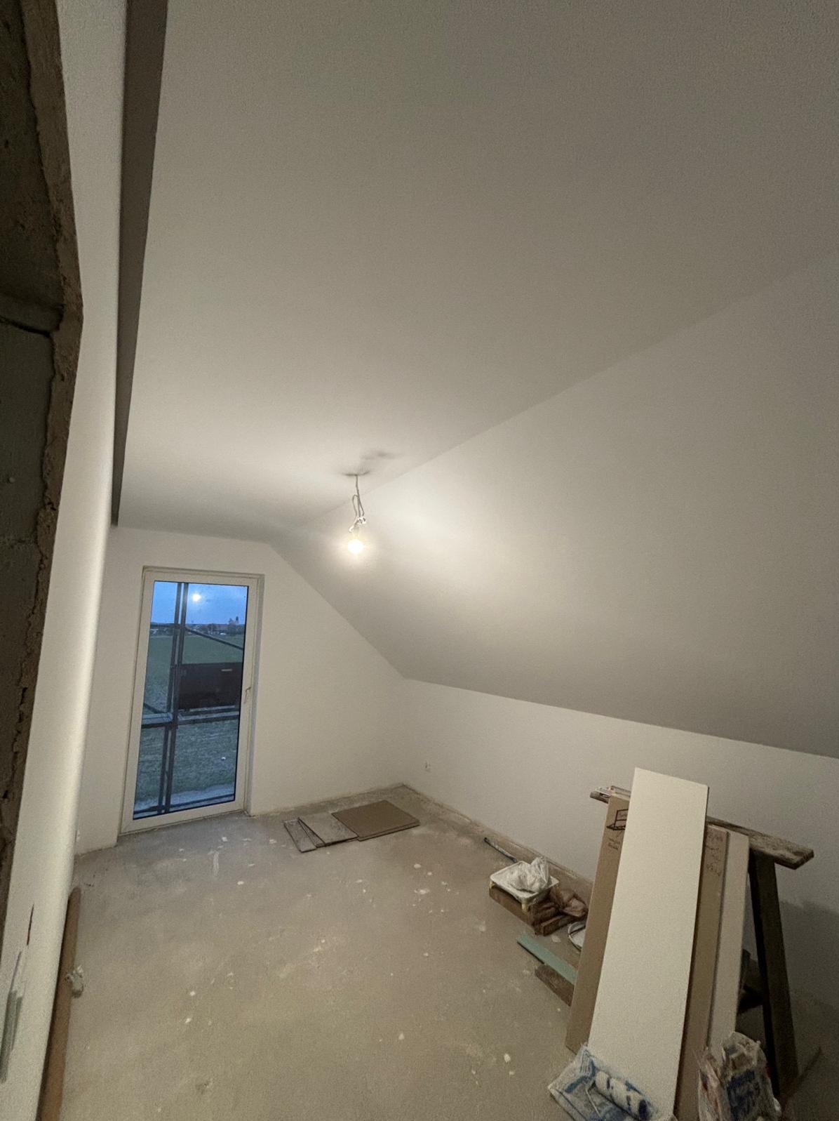 Poddasze po malowaniu na biało, widoczne skosy, okno w szczycie, sufit oświetlony żarówką. Na podłodze resztki materiałów budowlanych i narzędzia.