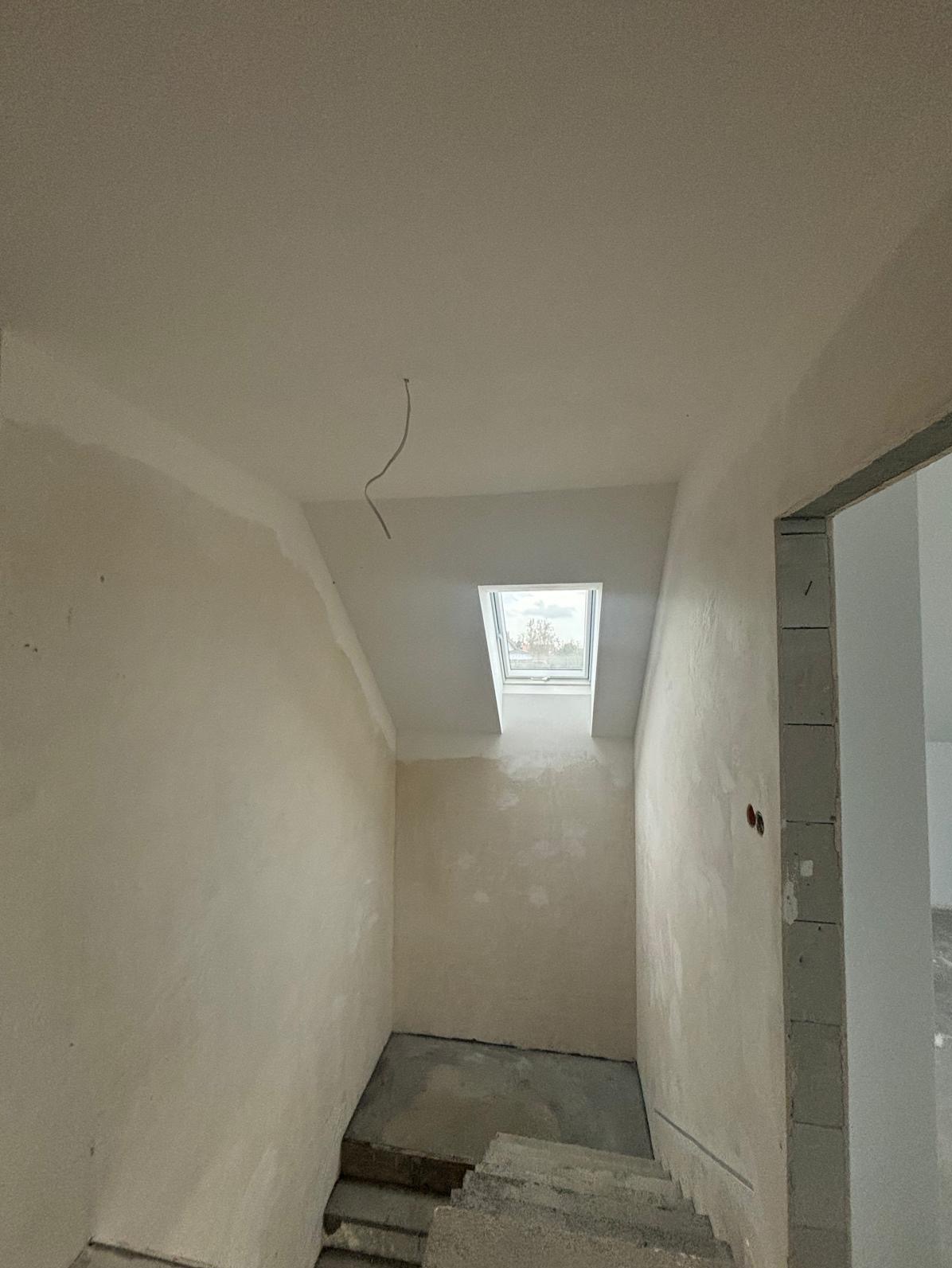 Wnętrze w trakcie remontu z oknem dachowym. Ściany po szpachlowaniu, ale przed malowaniem. Widoczne schody i wystający kabel elektryczny z sufitu.