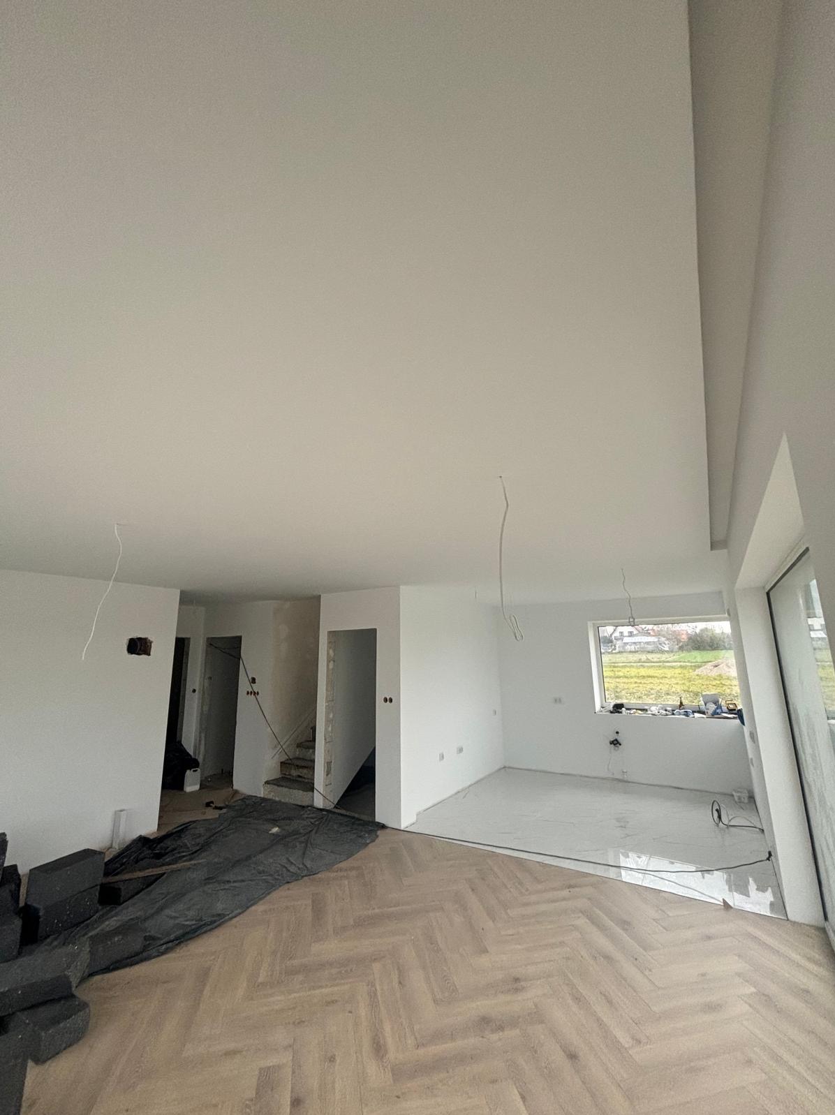 Jasne wnętrze w trakcie wykończenia: gładkie, białe ściany, parkiet w jodełkę, widoczne instalacje i okno z widokiem na zieleń. Częściowo zakryte podłoże.