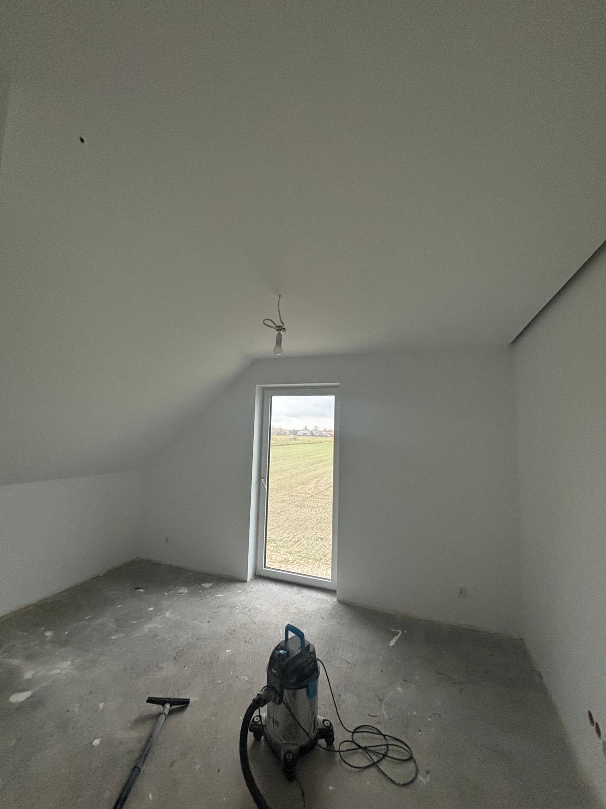 Poddasze po szpachlowaniu i malowaniu na biało, widoczny odkurzacz przemysłowy i niedokończona instalacja elektryczna. W tle widok z okna na pole.