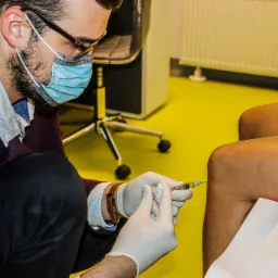 Lekarz w masce i rękawiczkach podaje zastrzyk w kolano pacjenta na żółtym tle gabinetu.