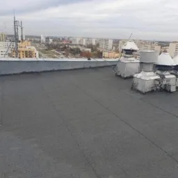 Płaski dach pokryty papą termozgrzewalną z widocznymi kominkami wentylacyjnymi na tle miejskiego krajobrazu w pochmurny dzień.