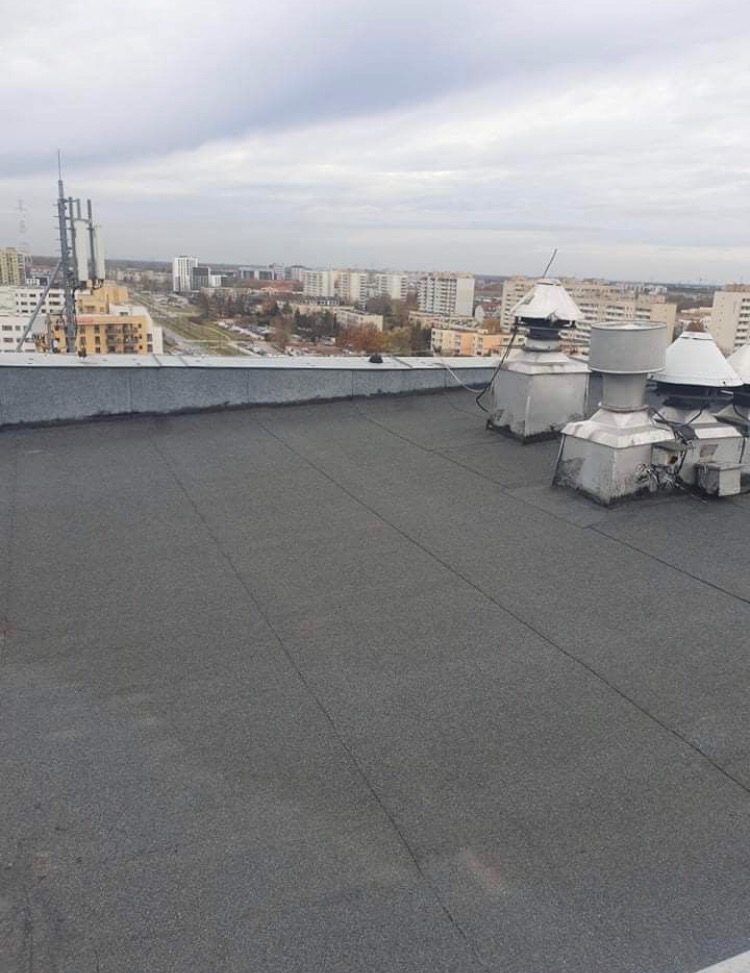 Płaski dach pokryty papą termozgrzewalną z widocznymi kominkami wentylacyjnymi na tle miejskiego krajobrazu w pochmurny dzień.