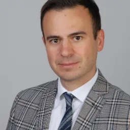 Portret mężczyzny w garniturze w kratę z niebieskim krawatem i wzorzystą poszetką w kieszonce, na szarym tle.