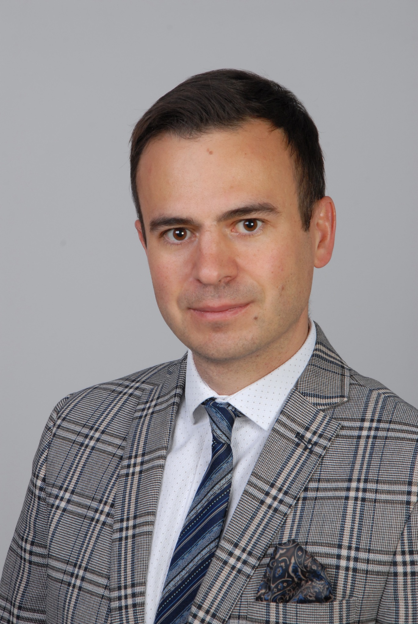 Portret mężczyzny w garniturze w kratę z niebieskim krawatem i wzorzystą poszetką w kieszonce, na szarym tle.