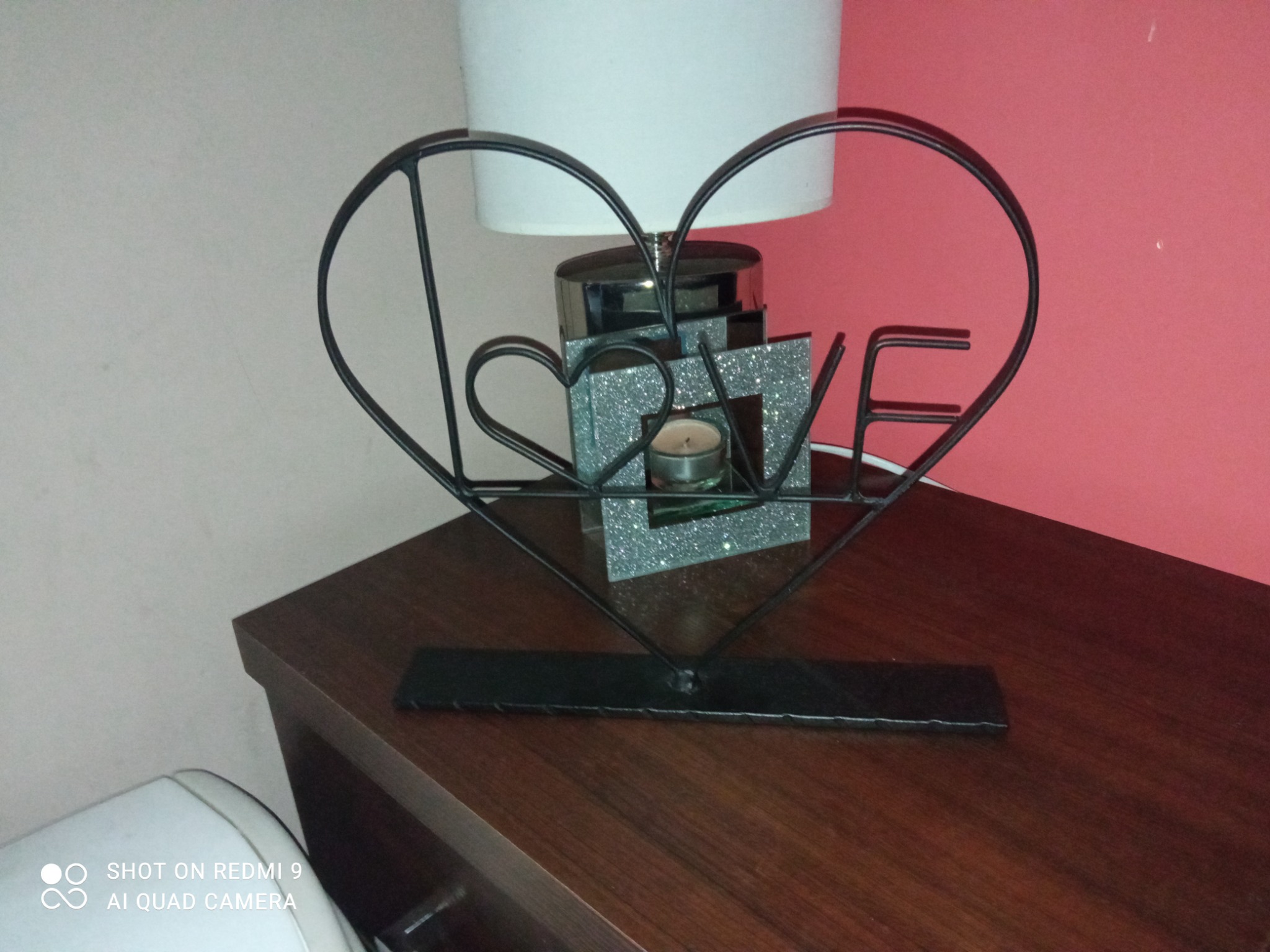 Metalowa dekoracja w kształcie serca z napisem LOVE, umieszczona na drewnianym blacie obok lampki nocnej.