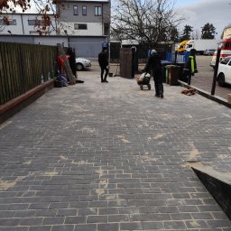 LexBud Oleksandr Pylypchuk - Ułożona szara kostka brukowa na podjeździe, z elementami piasku w spoinach. Pracownicy i sprzęt w tle sugerują trwające prace wykończeniowe.