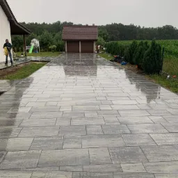 Ułożona, mokra kostka brukowa w odcieniach szarości prowadząca do garażu. W tle zieleń i dom z osobą z wężem ogrodowym. Widok na posesję w pochmurny dzień.