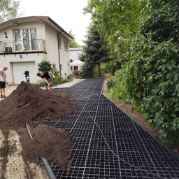 LexBud Oleksandr Pylypchuk - Przygotowanie podłoża: robotnicy rozgarniają ziemię na plastikowej siatce, tworząc podbudowę pod nawierzchnię na tle domu z garażem.