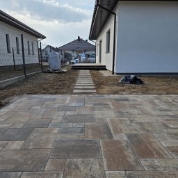 LexBud Oleksandr Pylypchuk - Ułożona kostka brukowa w odcieniach szarości i brązu prowadząca do domu. Widoczne worki z materiałem budowlanym i fragmenty ogrodu.